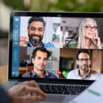 A person is engaged in a video conference on a laptop, which displays four participants in a grid format. The scene suggests remote implementation of a project, with visible charts and graphs on a document being held by the person.