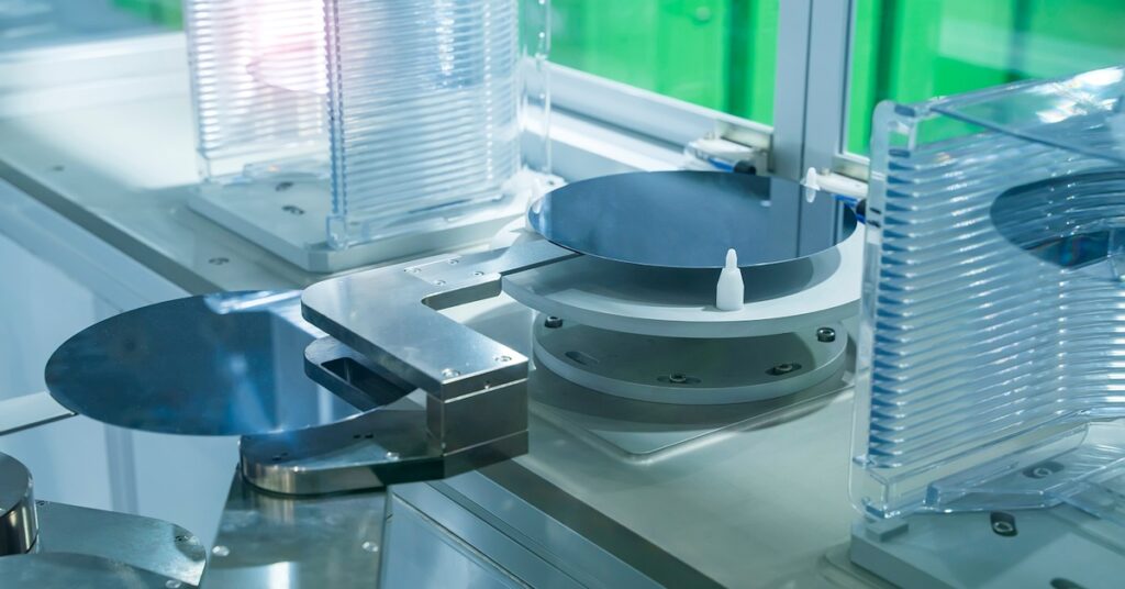 Close-up of silicon wafers placed on a mechanical stage in a cleanroom environment, surrounded by transparent containers and machinery components.