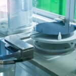 Close-up of silicon wafers placed on a mechanical stage in a cleanroom environment, surrounded by transparent containers and machinery components.