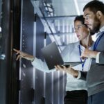 Two professionals analyzing data on a computer in a server room, focusing on advanced production planning and scheduling (APS)