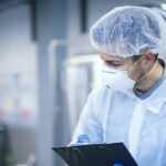 Technologist expert in protective uniform with hairnet and mask taking parameters from industrial machine in food production plant.