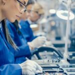 Female Electronics Factory Workers in Blue Work Coat and Protective Glasses Assembling Printed Circuit Boards for Smartphones with Tweezers. High Tech Factory with more Employees in the Background.