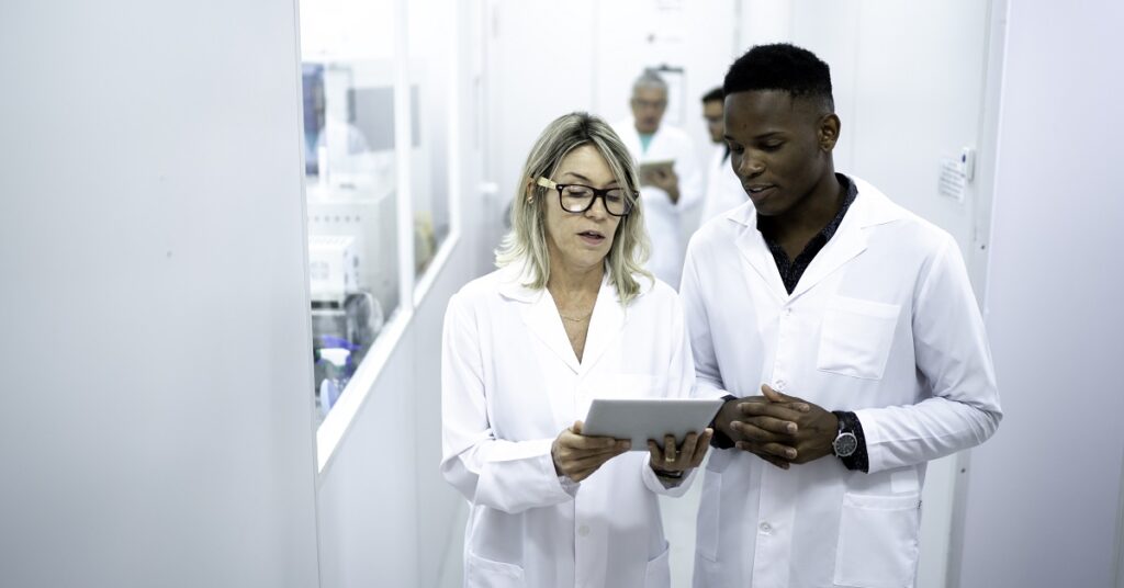 A man, with short black hair, and a woman, with blonde hair, in white lab coats, are standing in a brightly lit corridor, discussing data on a tablet in a laboratory setting while applying DataOps principles.
