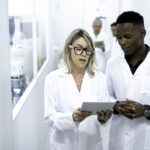 A man, with short black hair, and a woman, with blonde hair, in white lab coats, are standing in a brightly lit corridor, discussing data on a tablet in a laboratory setting while applying DataOps principles.