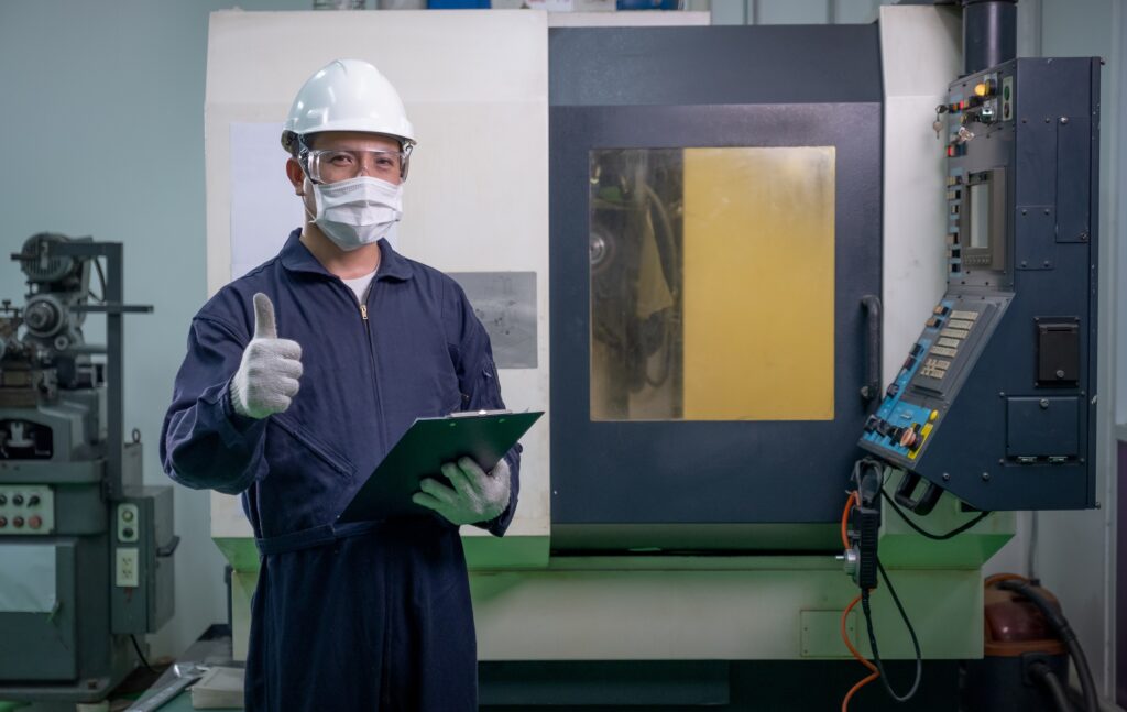 Factory worker man with mask and safety uniform hold the document also shows thumbs up and stand in front of operating machine