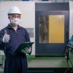 Factory worker man with mask and safety uniform hold the document also shows thumbs up and stand in front of operating machine