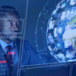 A man in a suit sitting in front of a large screen with a globe on it, with a serious expression on his face. The globe is surrounded by multiple screens, each displaying a different image of the world, and, on the top left corner, it has “Stages”, which may highlight the stages of the multisite MES project.