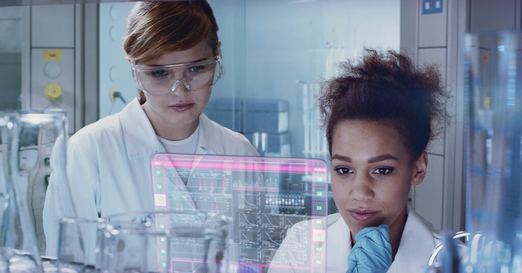 Two young women, wearing white lab coats and safety goggles, standing in front of a large computer screen. In the background, there are several test tubes and other medical devices.