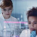 Two young women, wearing white lab coats and safety goggles, standing in front of a large computer screen. In the background, there are several test tubes and other medical devices.